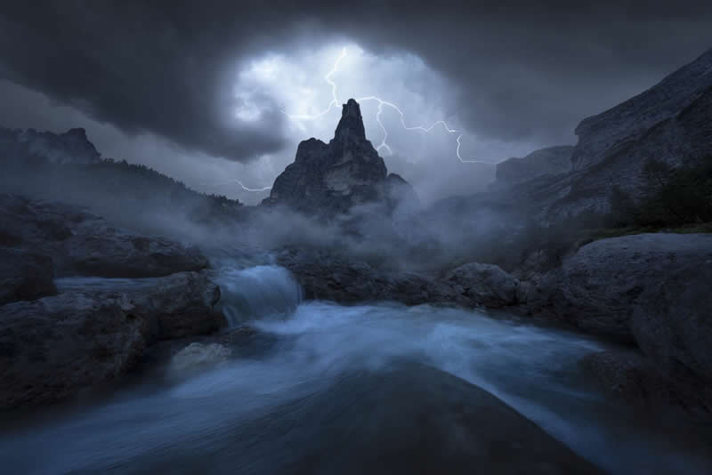 2026 Memorial Maria Luisa Awards: Best Mountain & Nature Photos Revealed 37 Lightning striking behind a rocky mountain peak with mist, flowing water, and dark storm clouds creating a dramatic landscape.