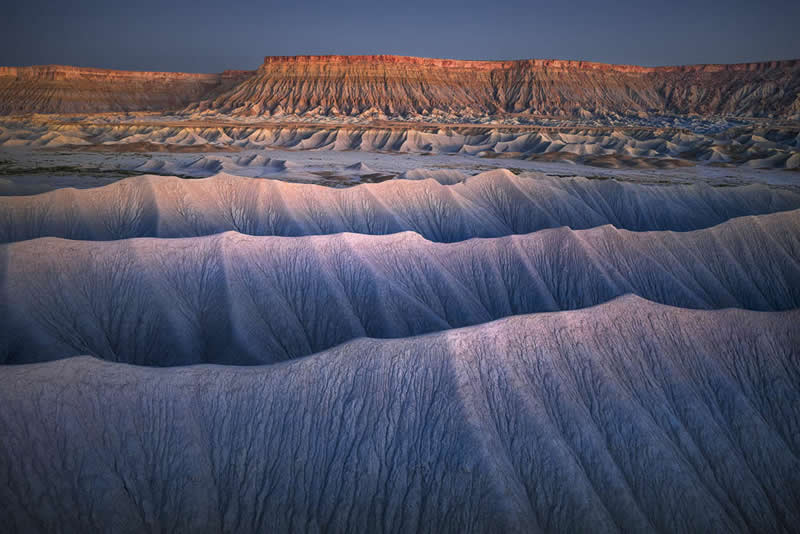 2026 Memorial Maria Luisa Awards: Best Mountain & Nature Photos Revealed 58 Eroded badlands with wave-like ridges in the foreground and a sunlit cliff in the background under soft evening light.