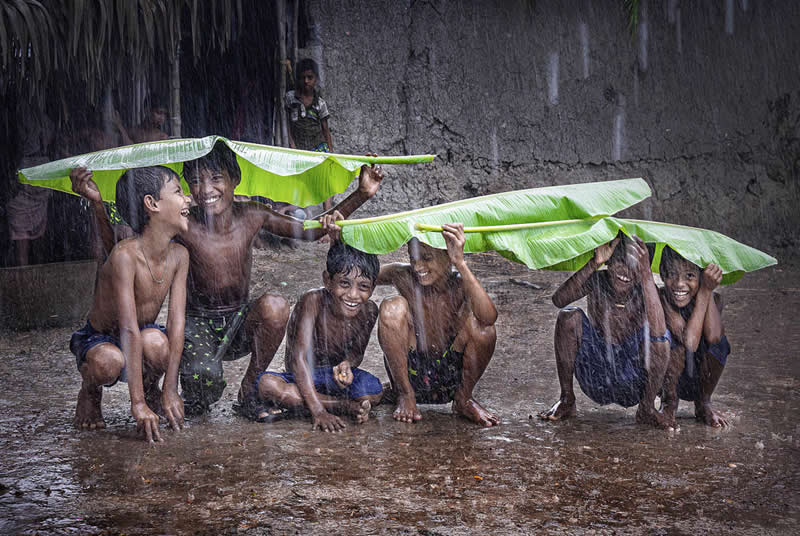 2026 Memorial Maria Luisa Awards: Best Mountain & Nature Photos Revealed 64 Children playing in heavy rain, using banana leaves as umbrellas, smiling and crouching together in a muddy rural setting.