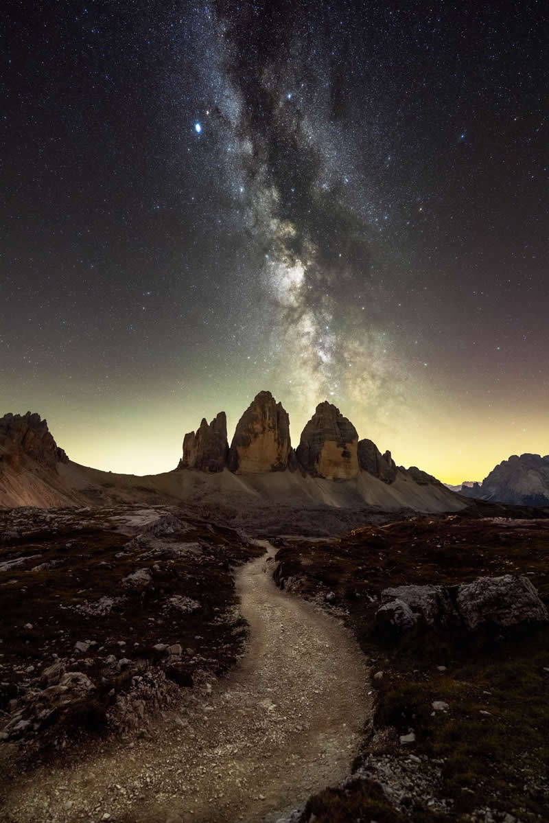 The Milky Way stretches above jagged mountain peaks, while a winding trail cuts through the landscape below, all beneath a sky bursting with stars.