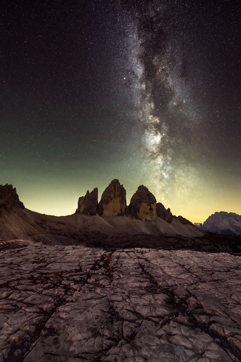 Milky Way galaxy rising above dramatic rocky mountain peaks with textured stone foreground under a clear star-filled night sky.