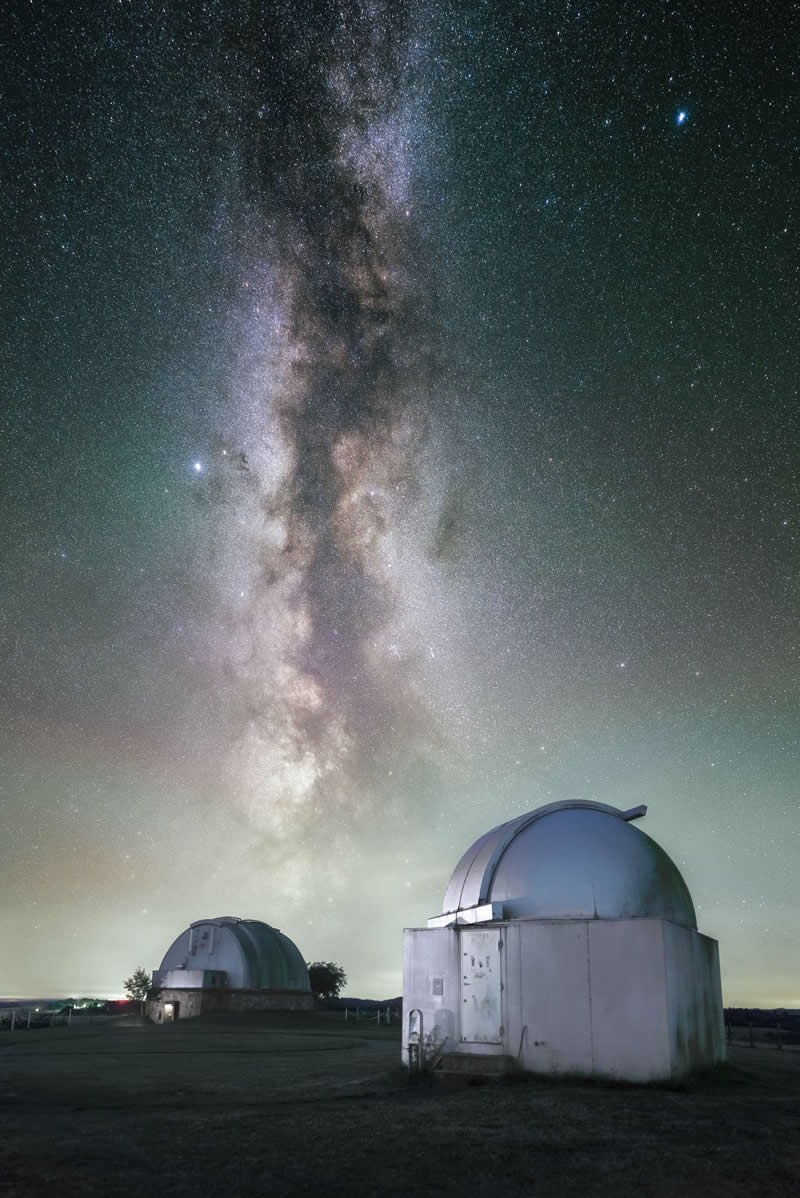 Astronomical observatories beneath the Milky Way galaxy with thousands of stars visible in the night sky above the domed telescope buildings.