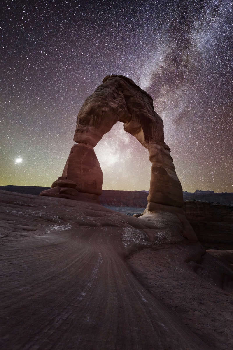 Natural sandstone arch under the Milky Way galaxy with thousands of stars shining above a desert landscape at night.
