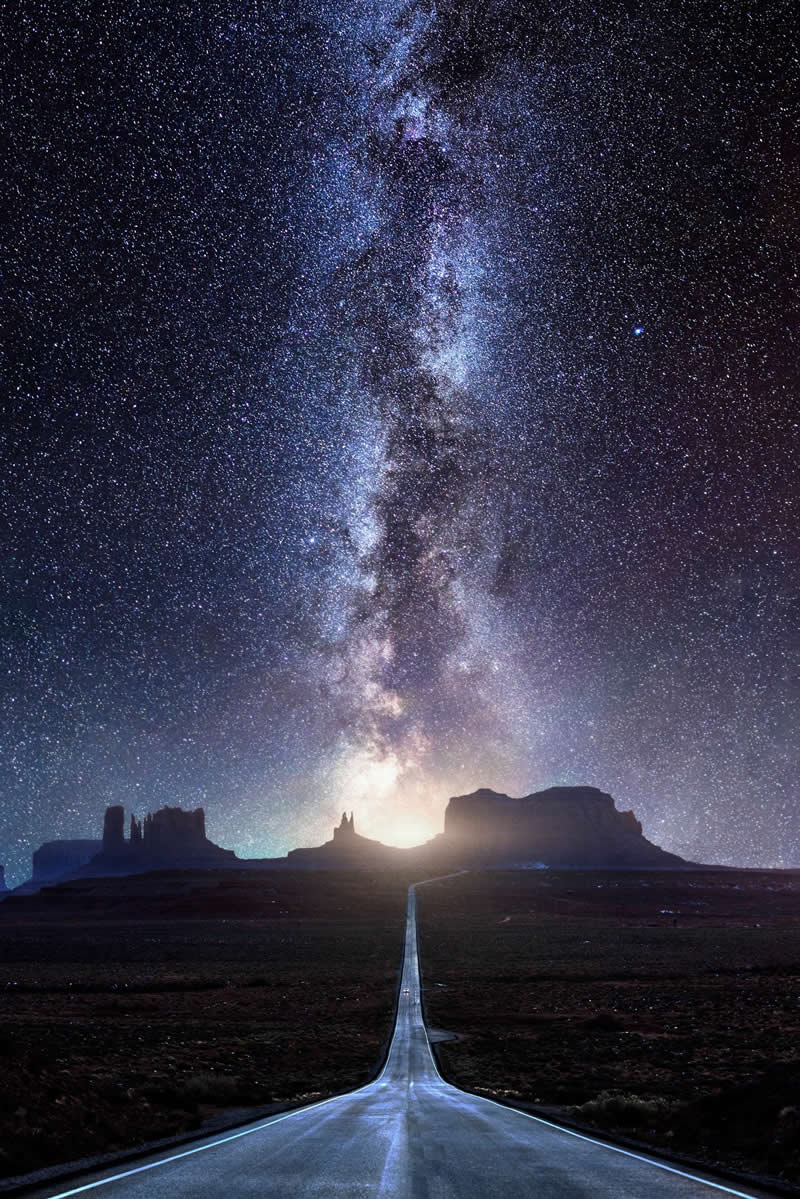 Long straight desert road leading toward distant mesas beneath a bright Milky Way galaxy filled with thousands of stars in the night sky.