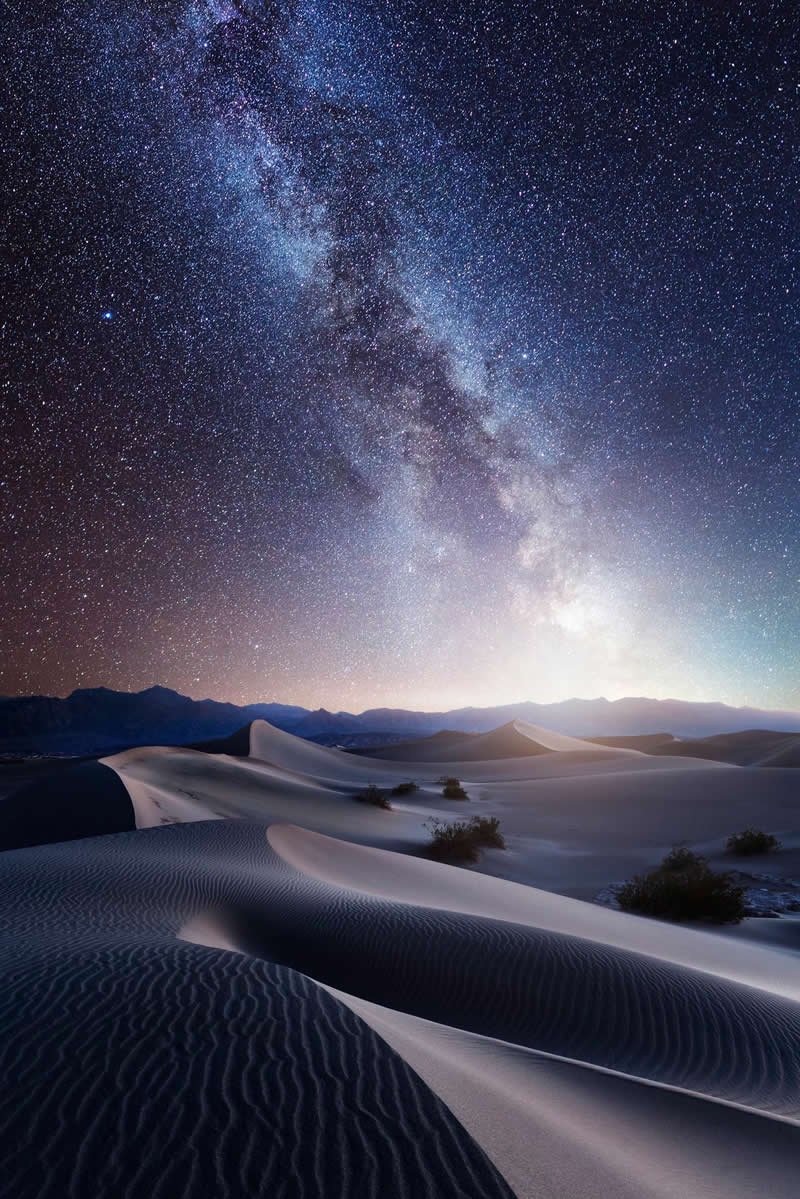 Milky Way galaxy shining above smooth desert sand dunes with mountains in the distance under a clear star-filled night sky.