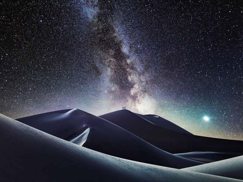 Two people standing on sand dunes under a bright Milky Way galaxy with countless stars filling the night sky above the desert landscape.