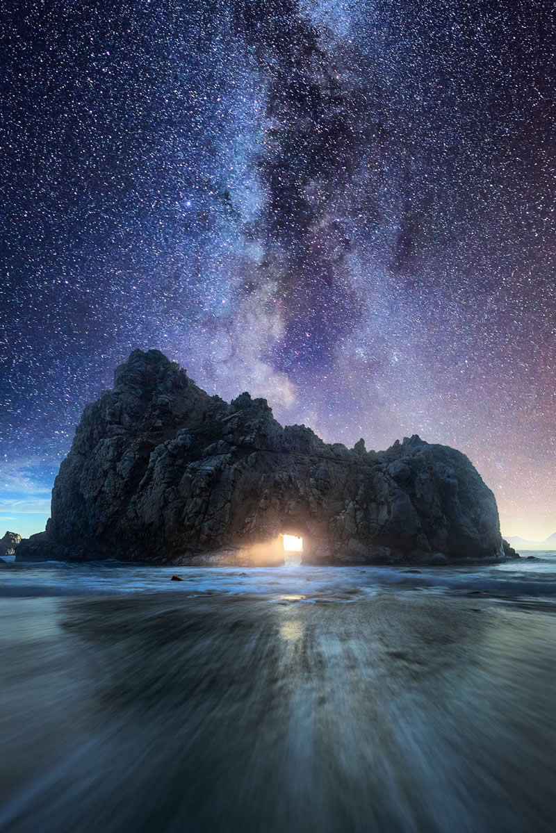 Milky Way galaxy rising above a rocky sea arch with light glowing through the opening while waves blur across the shoreline at night.