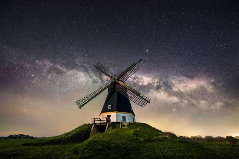 Traditional windmill on a grassy hill beneath the Milky Way galaxy stretching across a clear star-filled night sky.
