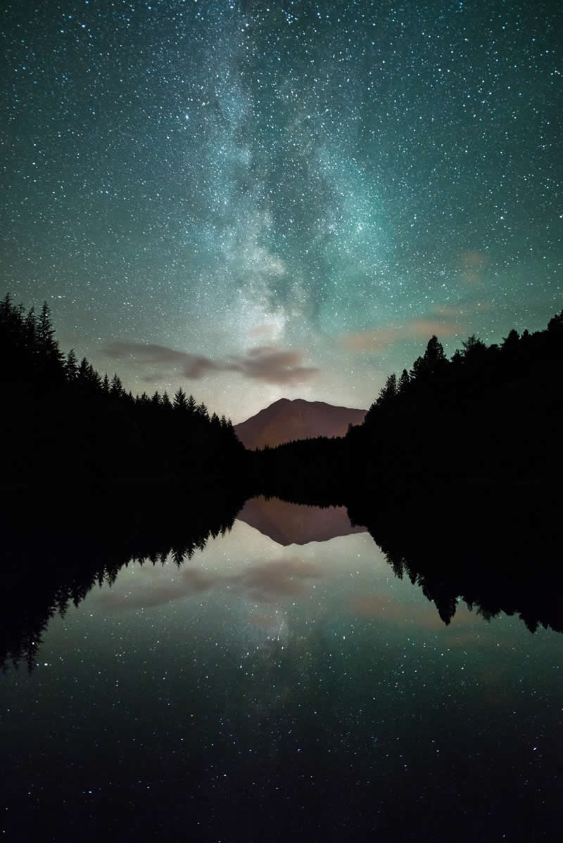 Milky Way galaxy reflected in a calm mountain lake surrounded by dark forest silhouettes with a distant mountain under a star-filled night sky.