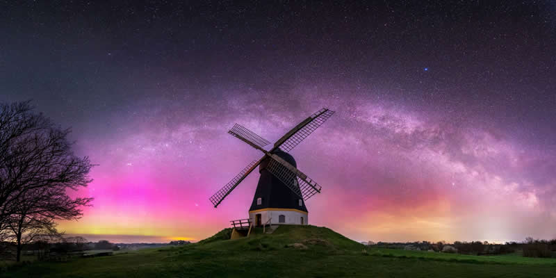 Windmill on a grassy hill under a colorful Milky Way arch with pink and purple night sky glowing above the landscape.