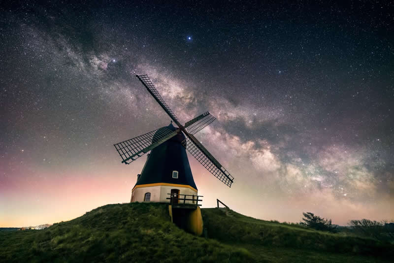 Traditional windmill on a grassy hill under a glowing Milky Way galaxy with stars stretching across the night sky.