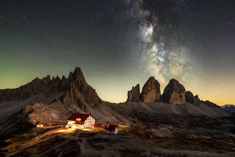 A glowing mountain cabin sits below dramatic rocky peaks, while the Milky Way arcs across a crisp, clear night sky above.