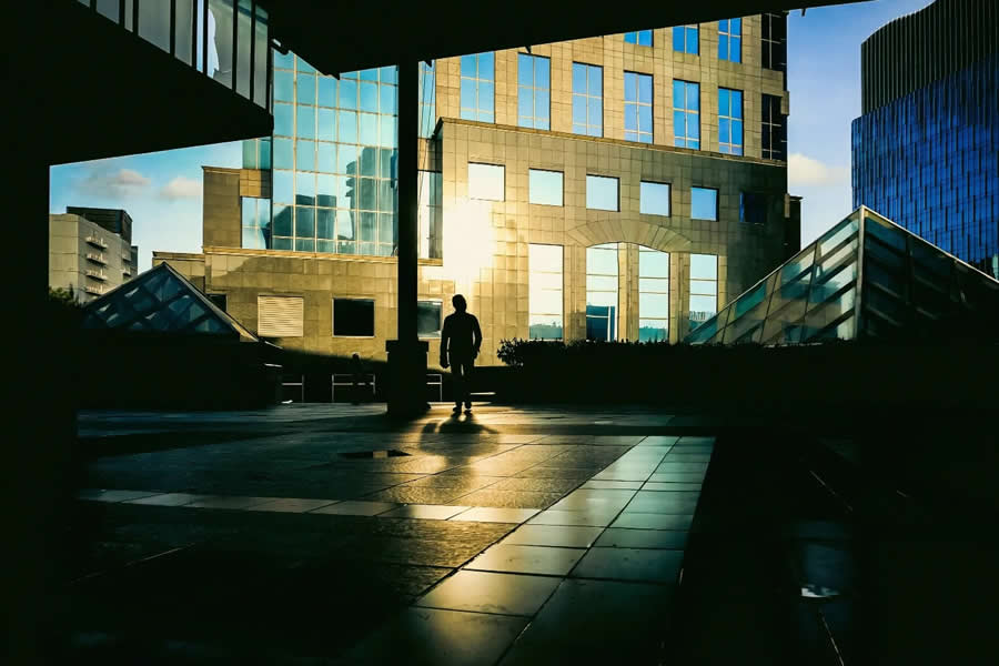 Silhouette of a person standing in front of modern glass buildings with golden sunlight reflections, creating strong geometric lines and a cinematic urban street photography composition.