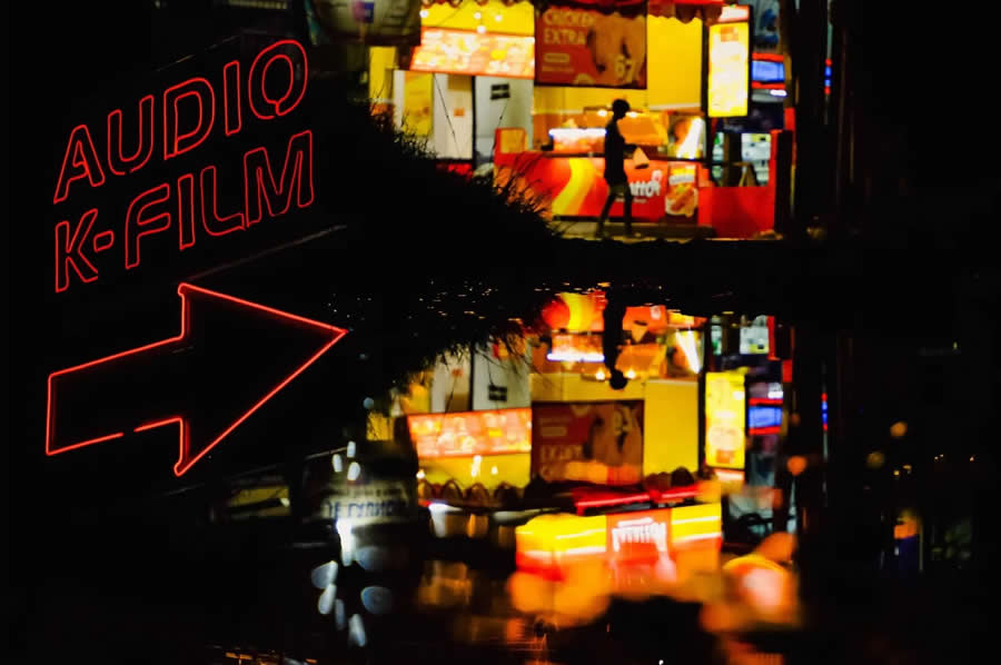 Neon sign reading “Audio K-Film” with bright street lights and a person walking, reflected in a wet surface, creating colorful reflections and a dynamic night street photography scene.