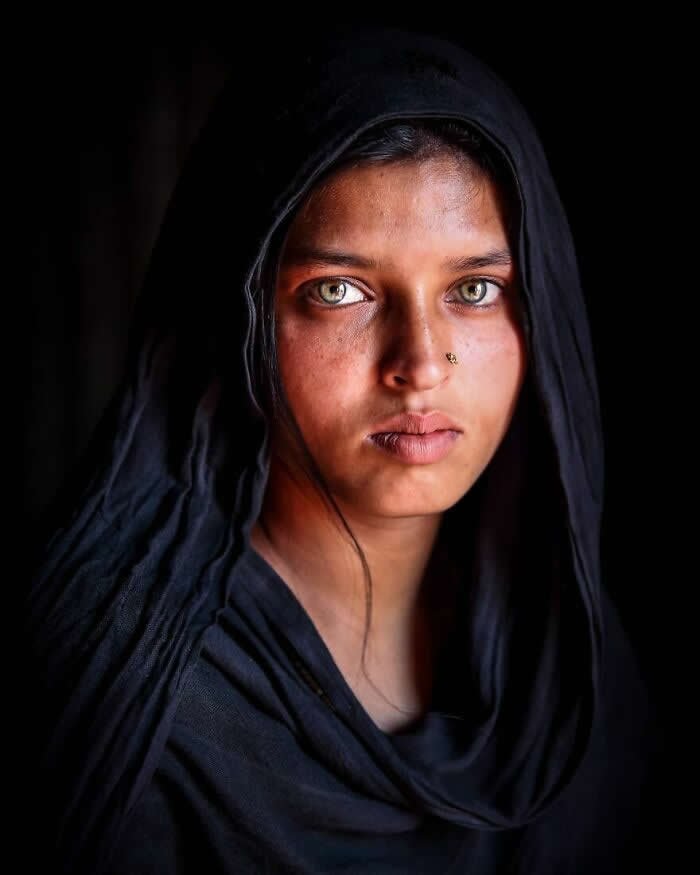 A soulful portrait of a young Bangladeshi woman wearing a black veil, her striking light green eyes glowing softly against a dark background, captured in dramatic natural light that highlights her calm yet powerful expression.