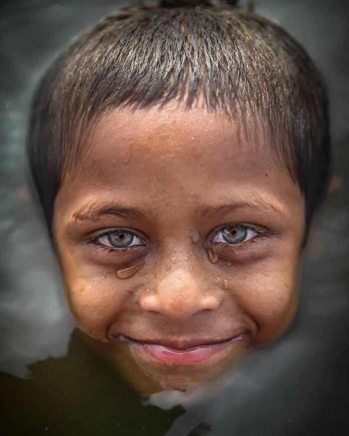 A close-up portrait of a Bangladeshi child with striking light green-gray eyes, smiling gently as tears run down their cheeks, captured in soft natural light with a blurred dark background that emphasizes the emotional expression.