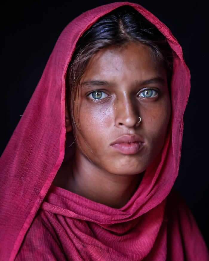 A close-up portrait of a young Bangladeshi woman wearing a pink headscarf, her striking light blue-green eyes gazing directly at the camera against a dark background, highlighting her calm expression and luminous gaze.