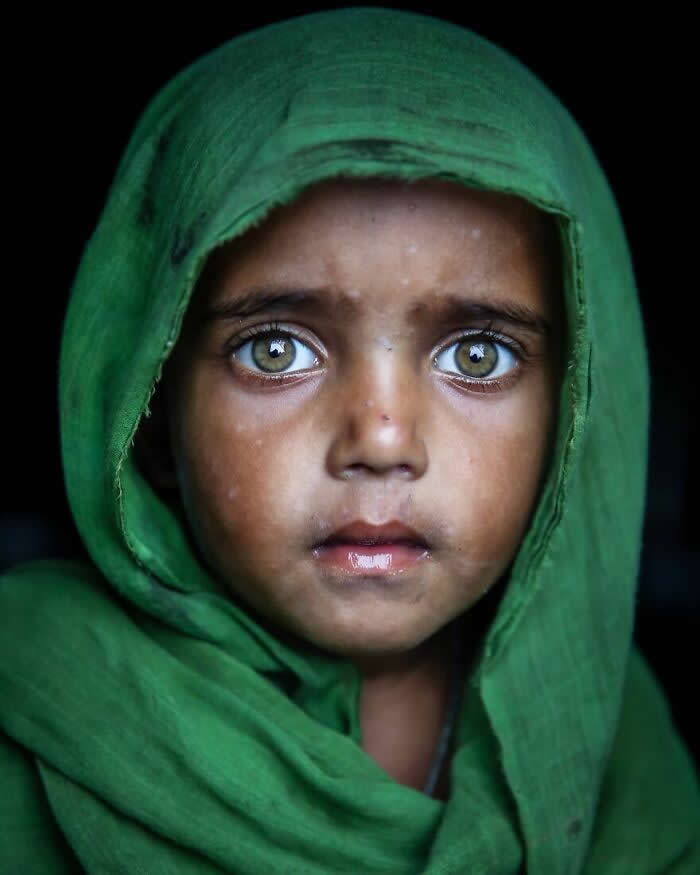 A close-up portrait of a young Bangladeshi child wearing a green headscarf, with striking light gray-green eyes gazing wide toward the camera against a dark background, capturing a tender and soulful expression.