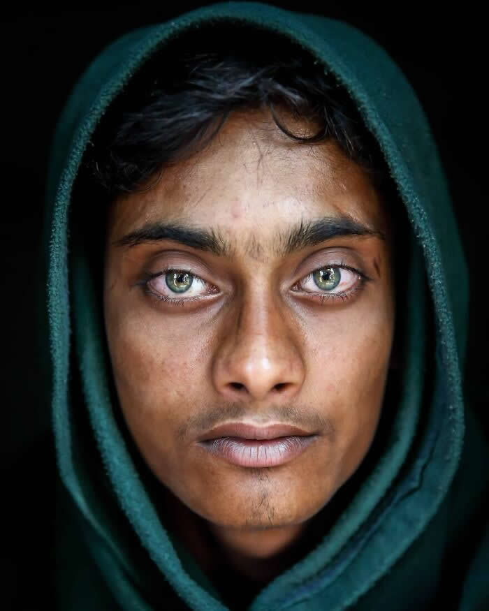 A close-up portrait of a Bangladeshi man wearing a dark green hooded garment, his striking light gray-green eyes gazing directly at the camera against a dark background, highlighting a calm, intense, and soulful expression.