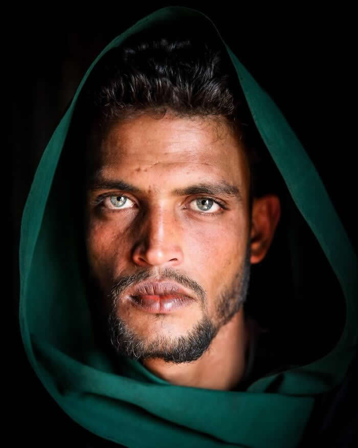 A close-up portrait of a Bangladeshi man wearing a deep green hooded scarf, his striking light gray-green eyes gazing intensely toward the camera against a dark background, highlighting rugged features and a powerful, soulful expression.