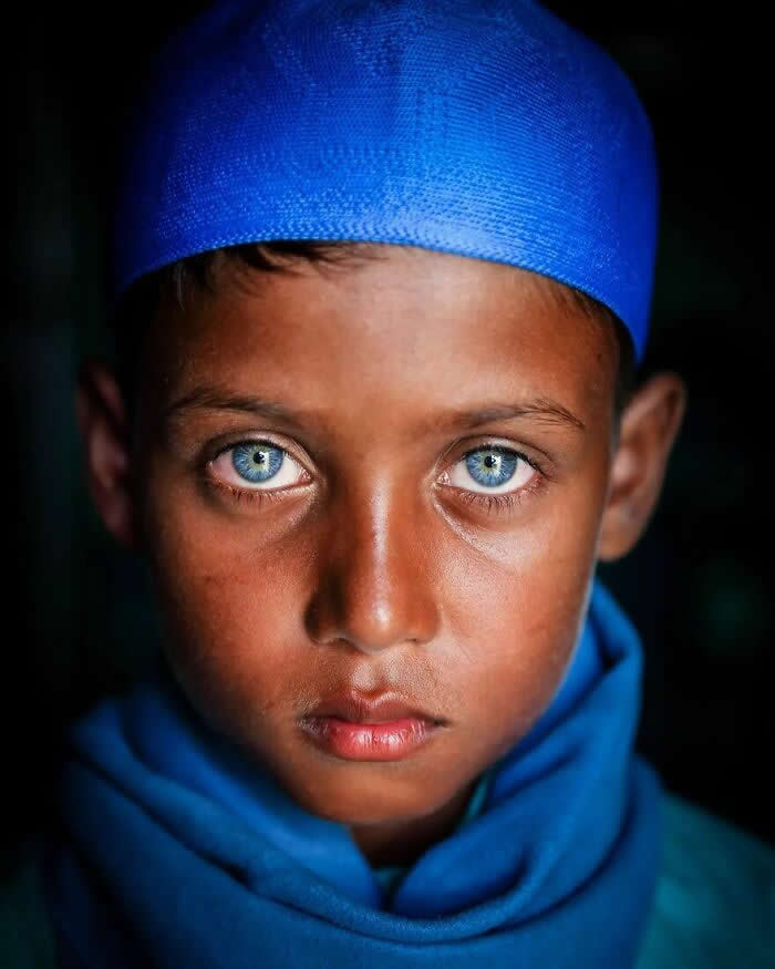 A close-up portrait of a Bangladeshi boy wearing a bright blue cap and scarf, with striking light blue eyes gazing directly at the camera against a dark background, highlighting his calm expression and luminous gaze.
