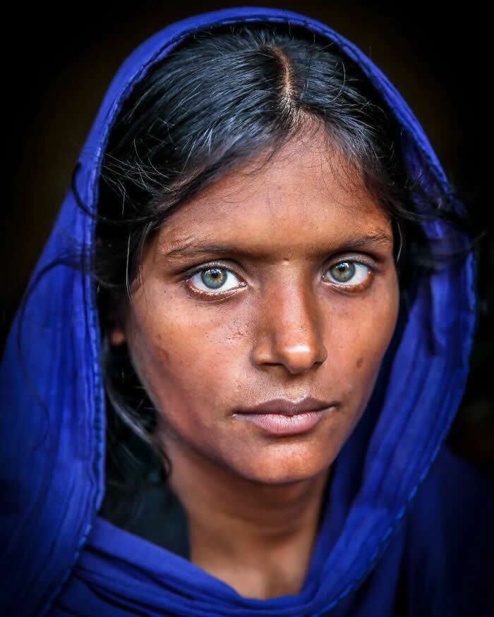 A close-up portrait of a Bangladeshi woman wearing a deep blue headscarf, her striking light blue-green eyes gazing calmly toward the camera against a dark background, highlighting a serene yet powerful expression.