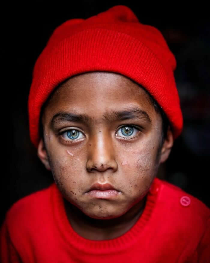 A close-up portrait of a Bangladeshi child wearing a bright red cap and sweater, with striking light blue-gray eyes gazing seriously toward the camera against a dark background, highlighting a powerful and emotional expression.