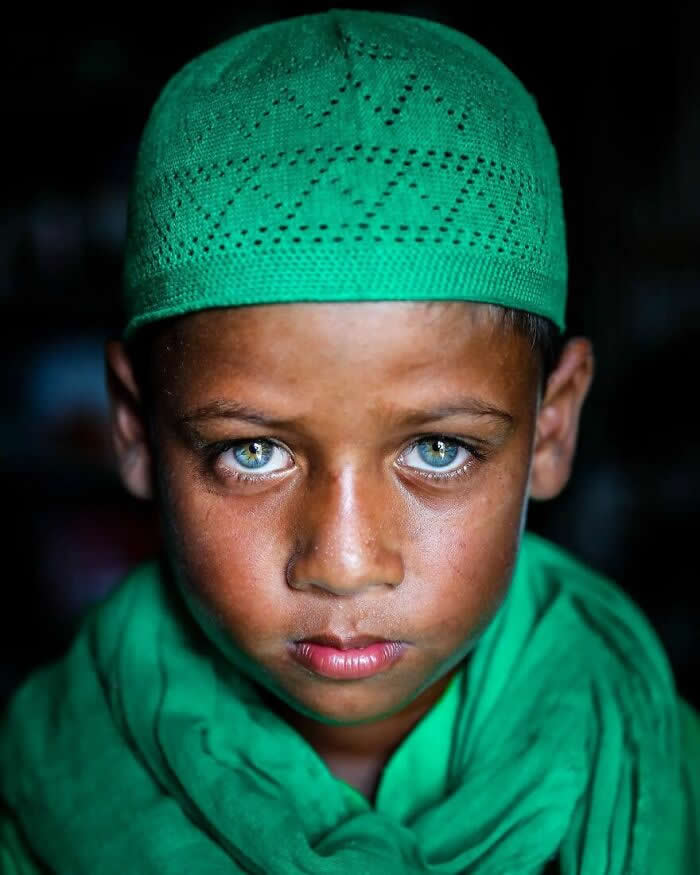 A close-up portrait of a Bangladeshi boy wearing a green prayer cap and scarf, his striking light blue-green eyes gazing seriously toward the camera against a dark background, highlighting a thoughtful and soulful expression.