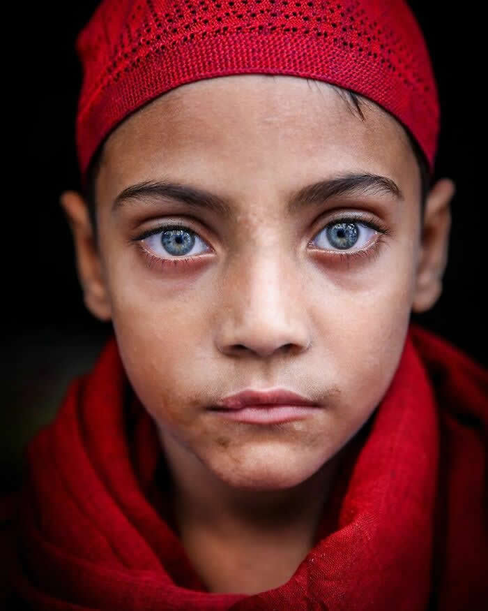 A close-up portrait of a Bangladeshi child wearing a red cap and scarf, with striking light blue eyes staring directly at the camera against a dark background, highlighting the child’s calm expression and luminous gaze.