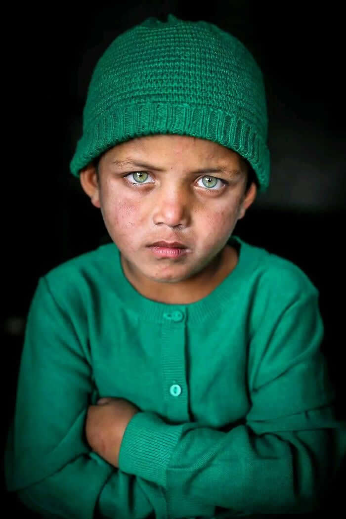 A close-up portrait of a Bangladeshi child wearing a green knit cap and sweater, with striking light blue-green eyes gazing intensely at the camera, arms crossed, against a dark background highlighting a powerful expression.