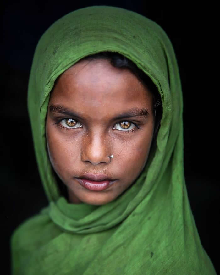 A close-up portrait of a young Bangladeshi girl wearing a green headscarf, her striking golden-brown eyes gazing intensely toward the camera against a dark background, highlighting her calm expression and luminous gaze.