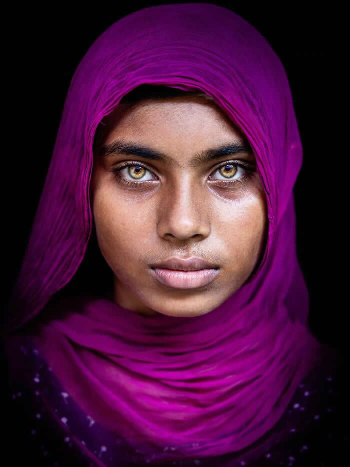 A close-up portrait of a young Bangladeshi girl wearing a deep magenta headscarf, her striking golden eyes glowing softly against a dark background, capturing a calm, soulful expression and powerful gaze.