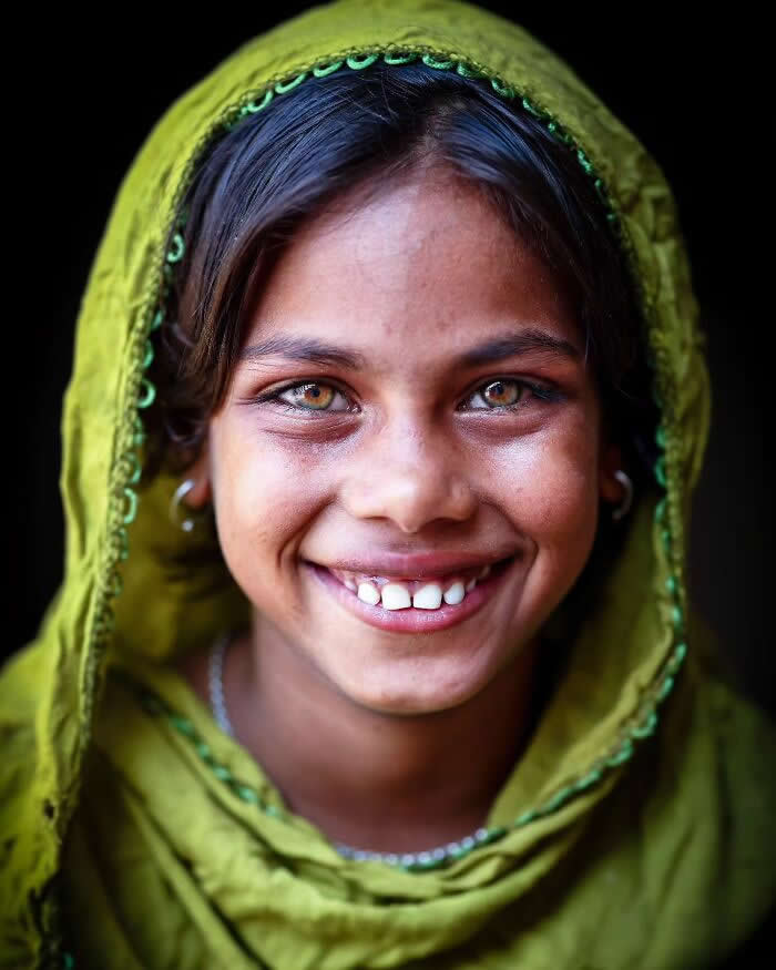 A close-up portrait of a smiling Bangladeshi girl wearing a green headscarf, her striking hazel eyes glowing warmly against a dark background, capturing a joyful expression and natural beauty.