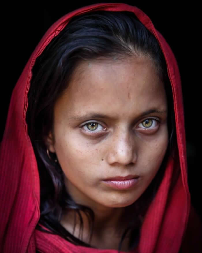 A close-up portrait of a young Bangladeshi girl wearing a red headscarf, her striking light green eyes gazing intensely toward the camera against a dark background, highlighting her calm yet powerful expression.