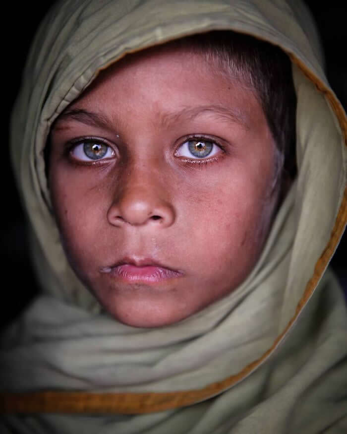 A close-up portrait of a Bangladeshi child wearing a beige headscarf, with striking light gray-green eyes gazing calmly toward the camera against a dark background, highlighting a thoughtful and soulful expression.