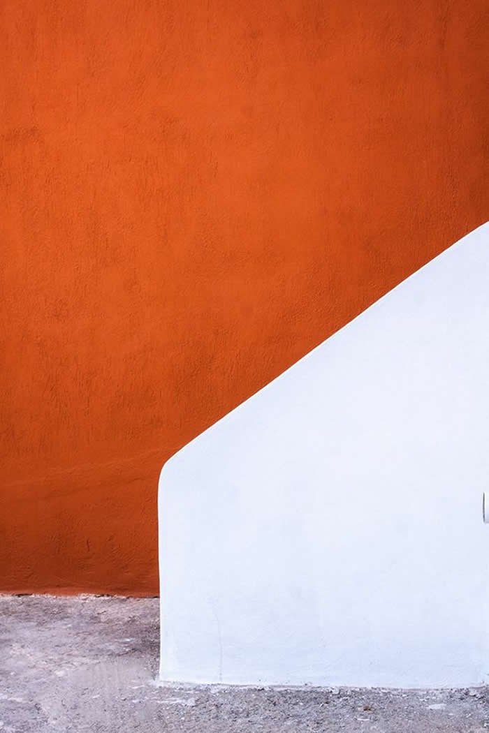 A minimalist composition of an orange textured wall intersected by a smooth white geometric shape, creating a clean diagonal contrast with subtle ground textures below.