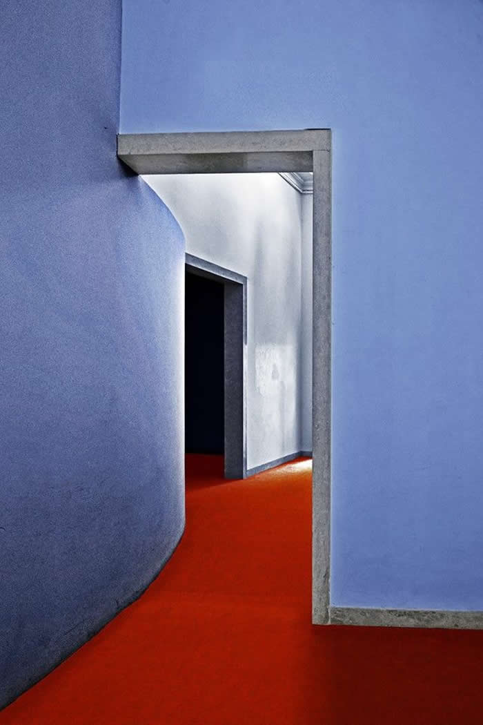 A minimalist interior scene with soft blue curved walls and sharp doorframes leading into a shadowy doorway, contrasted by a vivid red floor that creates depth and geometric flow.