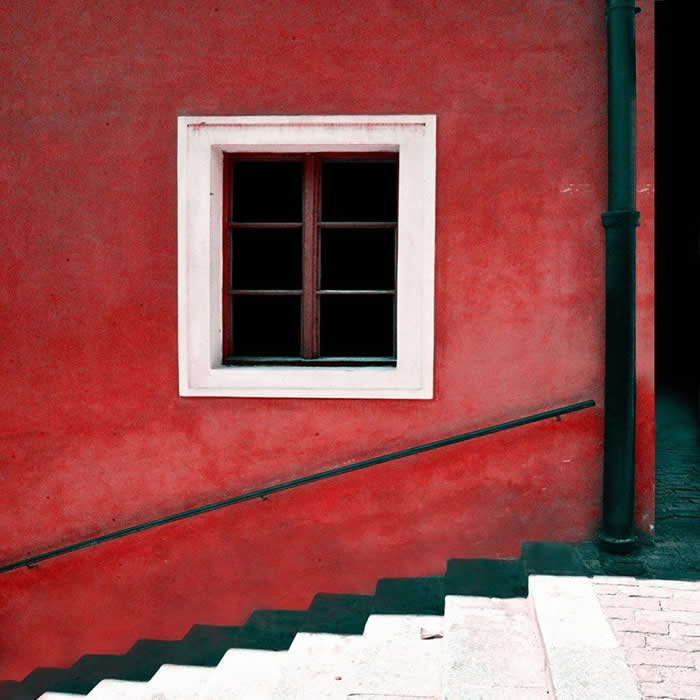 A minimalist scene featuring a red textured wall with a small square window, intersected by a diagonal staircase and railing, creating strong geometric lines and contrast.