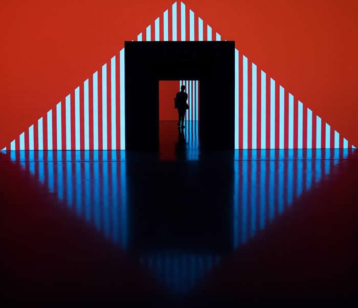 A silhouette of a person standing in a doorway within a bold geometric scene of red and blue striped triangular patterns, reflected on a glossy floor, creating symmetry and depth.