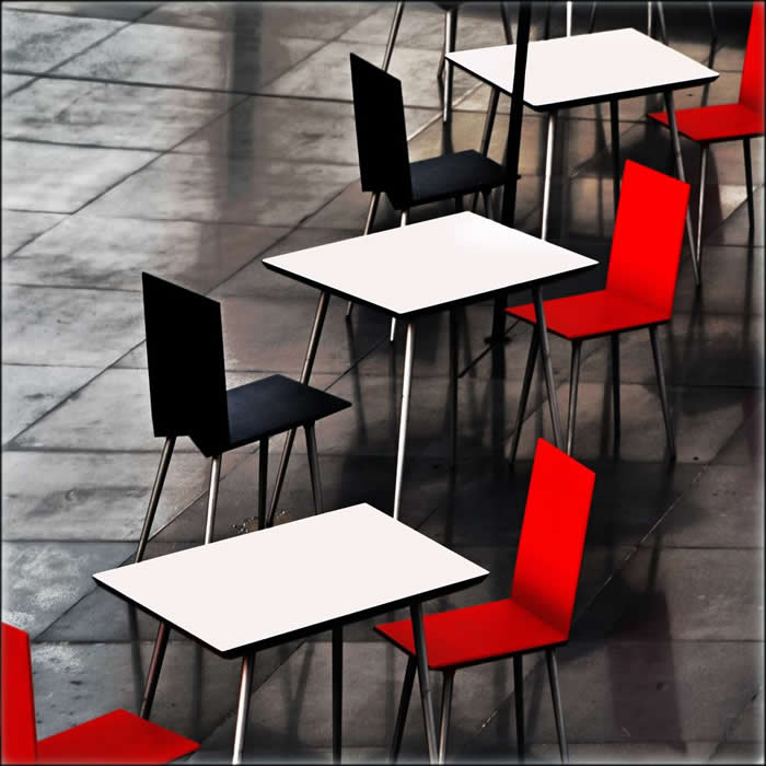 A minimalist arrangement of empty café tables with black and red chairs on a tiled floor, creating a rhythmic geometric pattern with strong contrast and repetition.