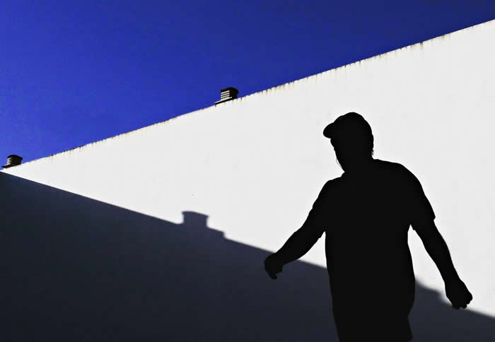 A silhouette of a person walking along a stark white wall under a deep blue sky, with sharp diagonal shadows creating a bold minimalist contrast and geometric composition.
