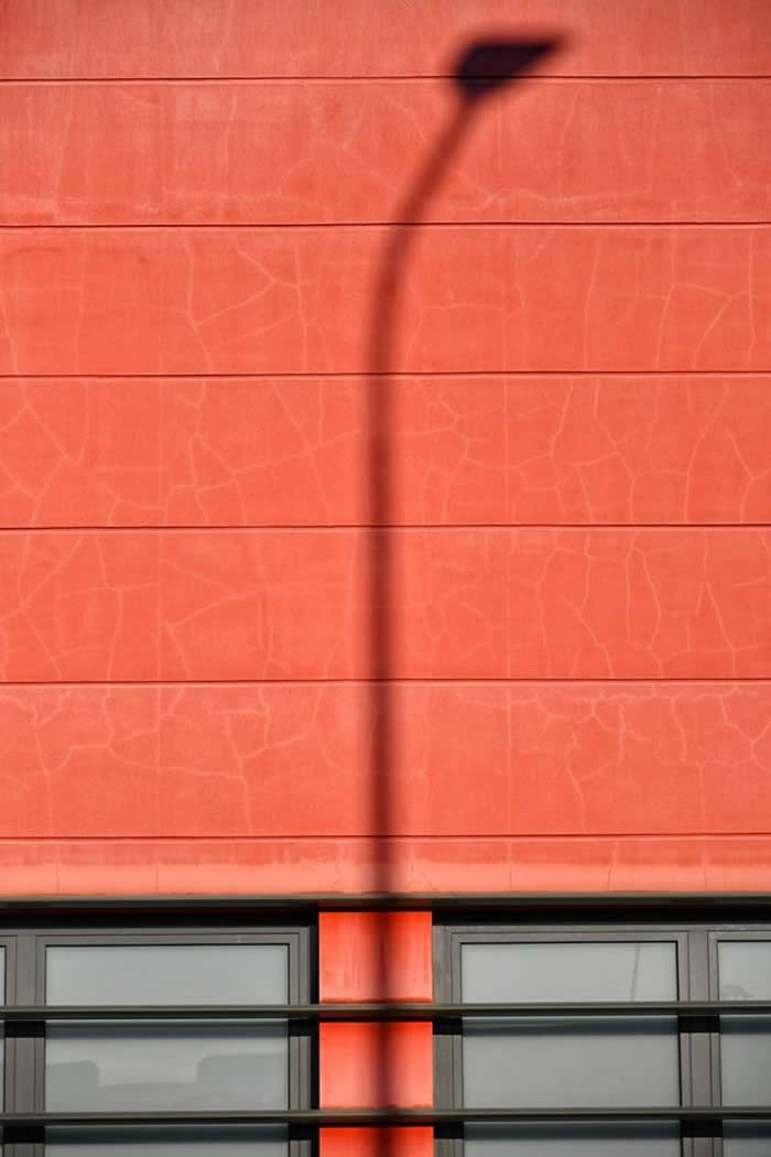 A minimalist composition featuring a textured red wall with horizontal lines, intersected by a tall, curved streetlight shadow, with a strip of windows at the bottom adding geometric contrast.