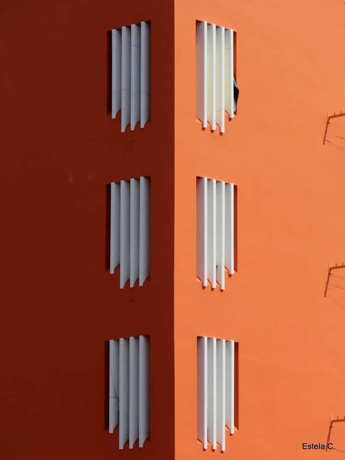 A minimalist architectural facade split into two shades of orange, featuring evenly spaced white window shutters that create a rhythmic pattern and balanced geometric symmetry.