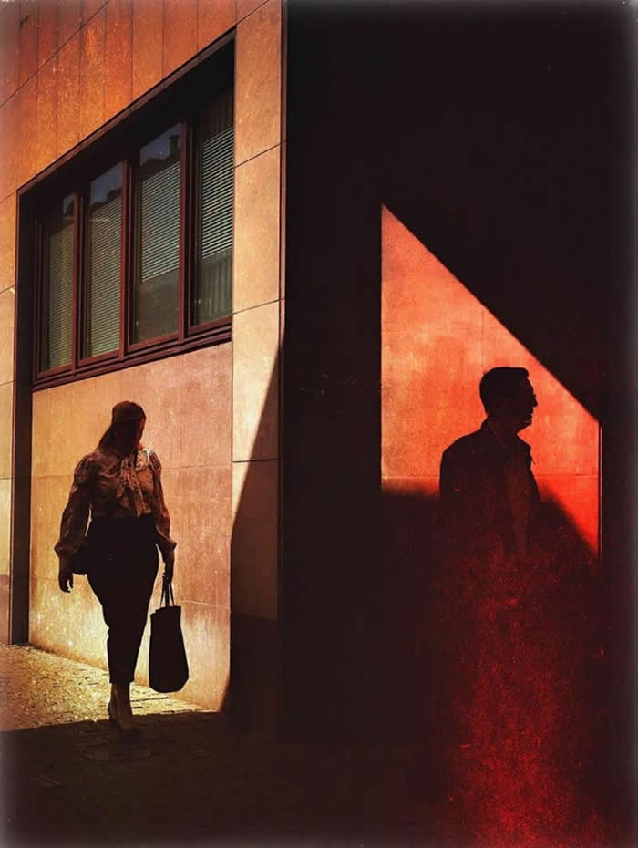 A dramatic street scene showing a woman walking in sunlight beside a building, while a man’s silhouette appears in a sharply lit red triangular shadow, creating contrast between light, shadow, and human presence.