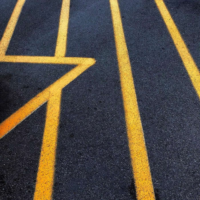 A minimalist view of dark asphalt marked with bold yellow road lines forming geometric patterns and directional shapes, creating strong contrast and abstract composition.