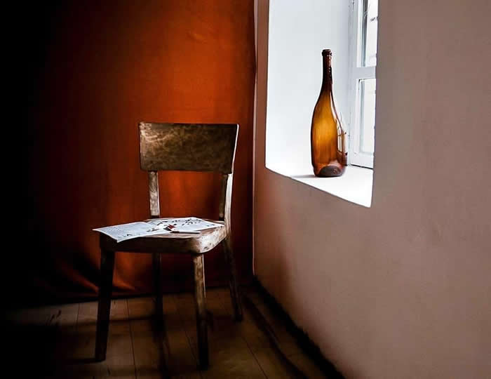 A quiet interior scene featuring a rustic wooden chair with scattered papers beside a window, where a brown glass bottle catches soft natural light against warm-toned walls.