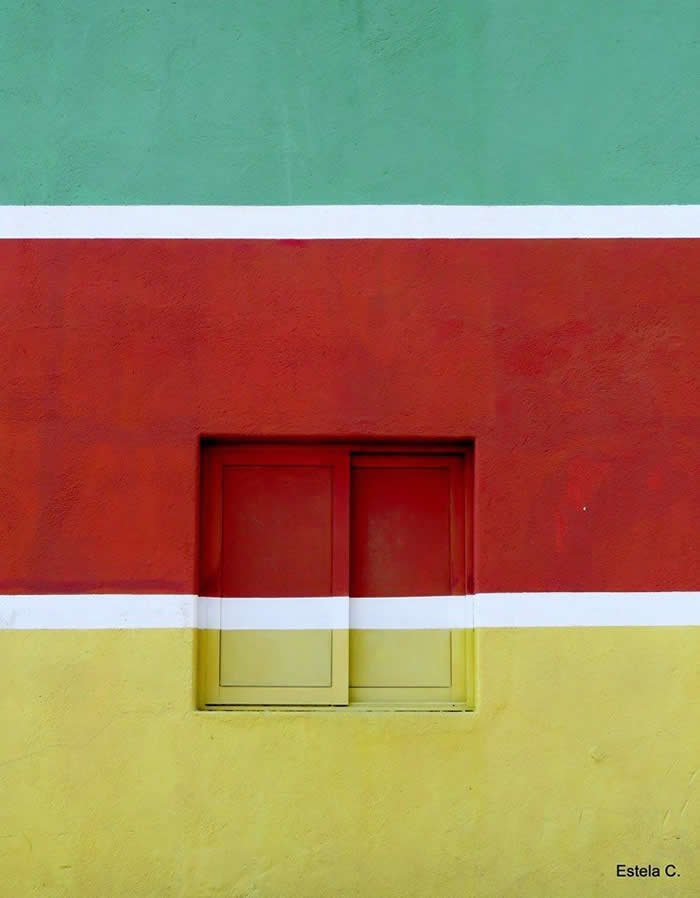 A minimalist composition of a colorful wall divided into horizontal bands of green, red, and yellow with white lines, centered by a small closed window blending into the geometric design.