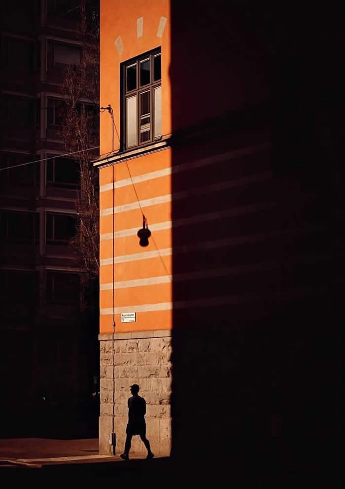 A lone person walking in shadow past a sunlit orange building with horizontal white stripes, where strong light and dark contrast create a dramatic geometric composition.
