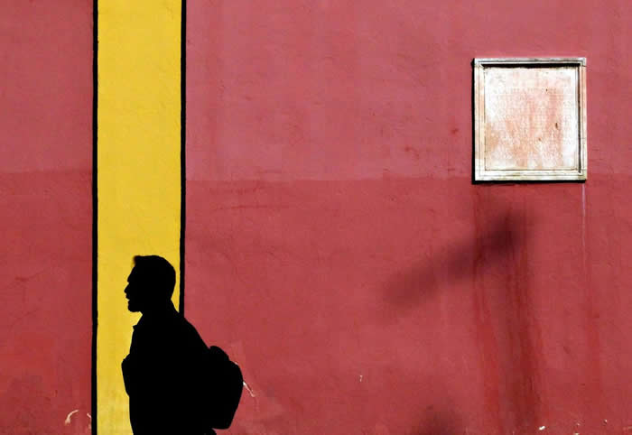 A silhouette of a person walking past a red wall with a bold vertical yellow stripe and a small window, creating a minimalist composition with strong contrast and geometric balance.