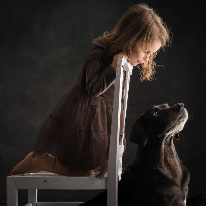 A young girl kneels on a chair, leaning forward as she gazes down at a black dog looking up at her, captured in soft studio lighting, expressing innocence, curiosity, and a gentle human–animal connection.